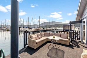 A covered outdoor seating area with a wicker sofa and table, overlooking a marina with boats.