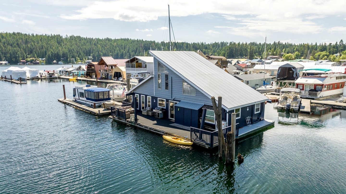 A waterfront house with a dock, a small boat, and a forested hill in the background.