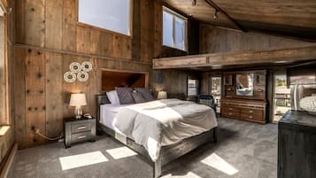 Une chambre avec un grand lit, une table de chevet, une commode et un miroir. Il y a une fenêtre au-dessus du lit et une porte qui donne sur une autre pièce.