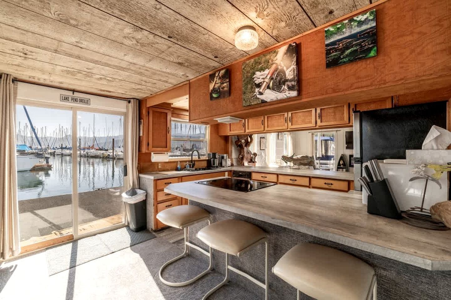 A modern kitchen with wooden cabinets, a large island with bar stools, and a view of a marina through a glass door.
