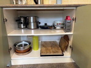 Ein Küchenschrank mit Edelstahl-Töpfen, einem Holzschneidebrett, einer Metallschüssel und einer gelben Wasserflasche.