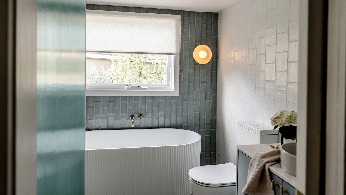 Une salle de bain moderne avec une baignoire sur pieds, une fenêtre avec un store blanc et un luminaire fixé au mur.