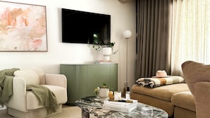 A living room with a sofa, a coffee table with books, a plant, and a television on the wall.