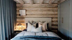 A bedroom with a wooden log wall, a bed with multiple pillows, a bedside table with a lamp, and a framed picture on the wall.