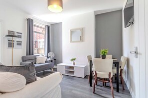 A modern living room with a sofa, a dining table with chairs, a small cabinet, and a large window with curtains. - Central 4-bed home in Kettering by Renzo, Ideal for Contractors, Close to Hospital (Kettering)