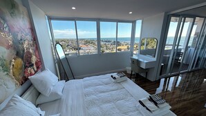 Ein Hotelzimmer mit einem großen Bett, einem Schreibtisch, einem Sessel und Blick auf Stadt und Meer.