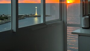 Ein Zimmer mit großen Fenstern, die Blick auf einen Sonnenuntergang über dem Meer bieten, mit einem Leuchtturm in der Ferne und einem Wasserkocher auf der Fensterbank.