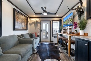 Ein modernes Interieur mit einer Couch, einem Kuhfellteppich, einem Deckenventilator und einer Küchenzeile mit Fernseher und Dekorationselementen.