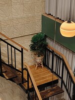 Una escalera con escalones de madera y barandillas negras, un atril tejido para plantas y una pared con textura y motivos de hojas.