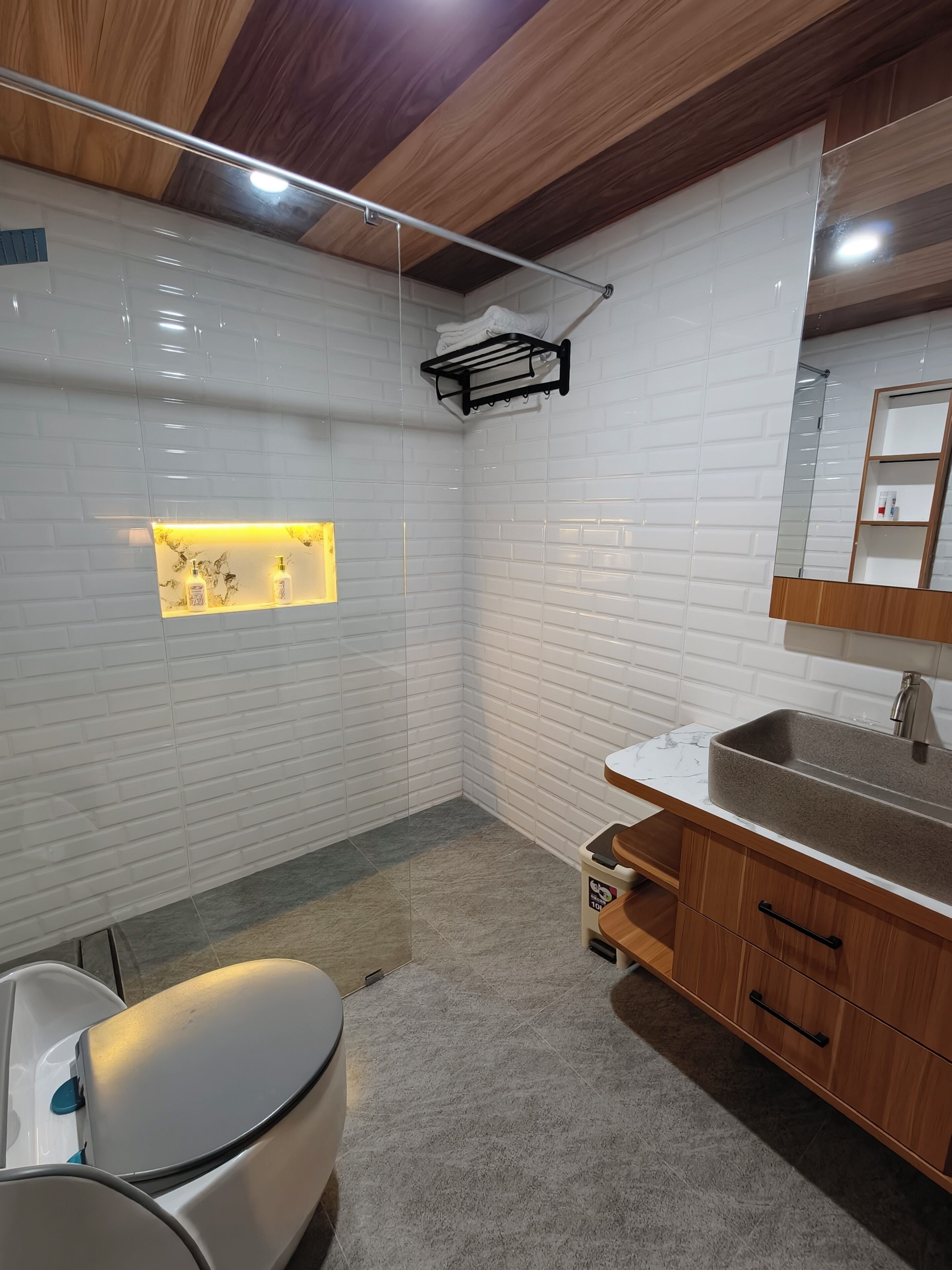 Un baño moderno con azulejos blancos tipo metro, lavamanos con encimera de mármol, armarios de madera y un estante empotrado con artículos de aseo.