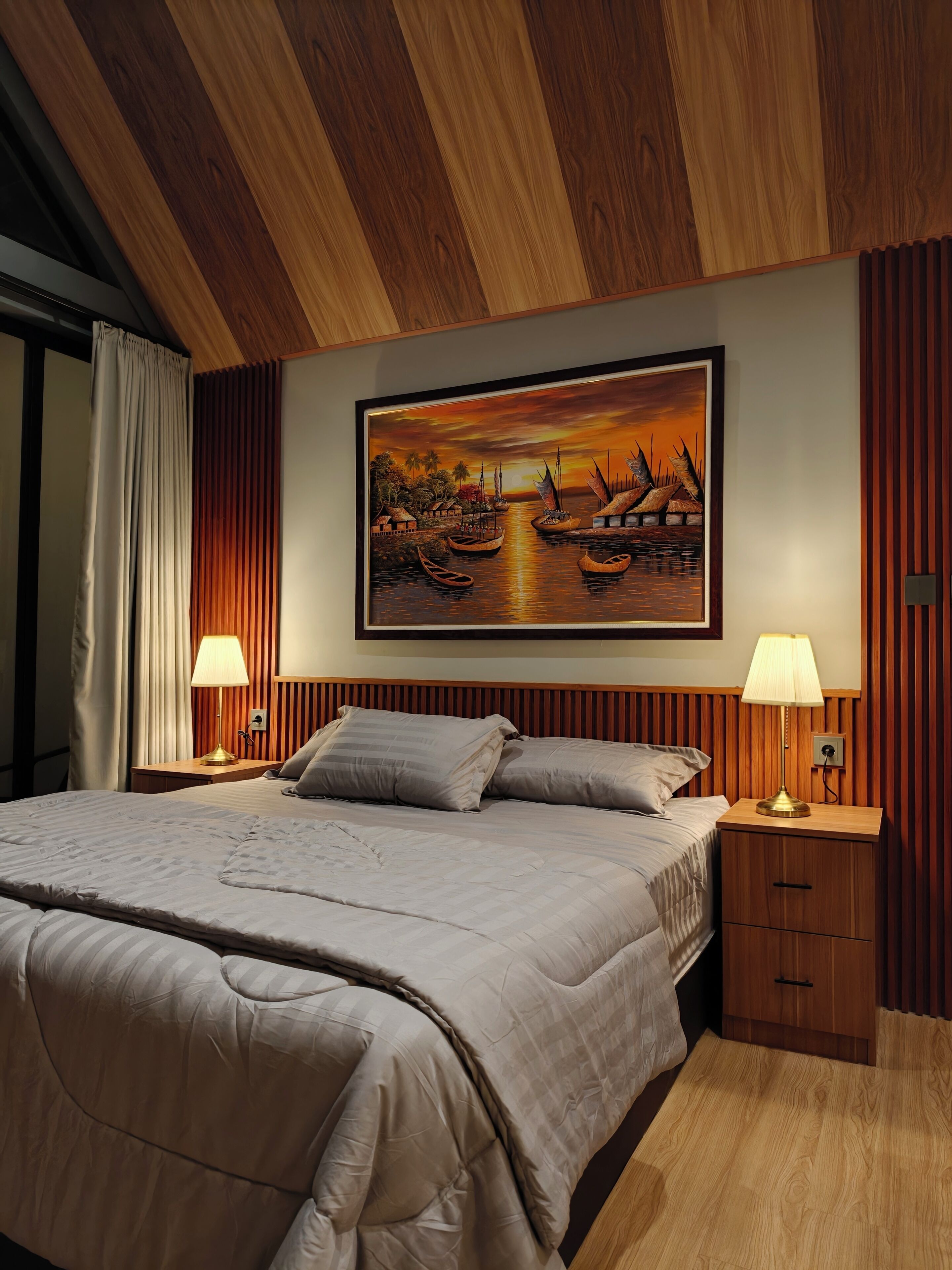 Ein Schlafzimmer mit einem Bett, zwei Nachttischlampen, einem Nachttisch, einem Holz-Kopfteil und einem Gemälde, das einen Sonnenuntergang über Wasser zeigt.