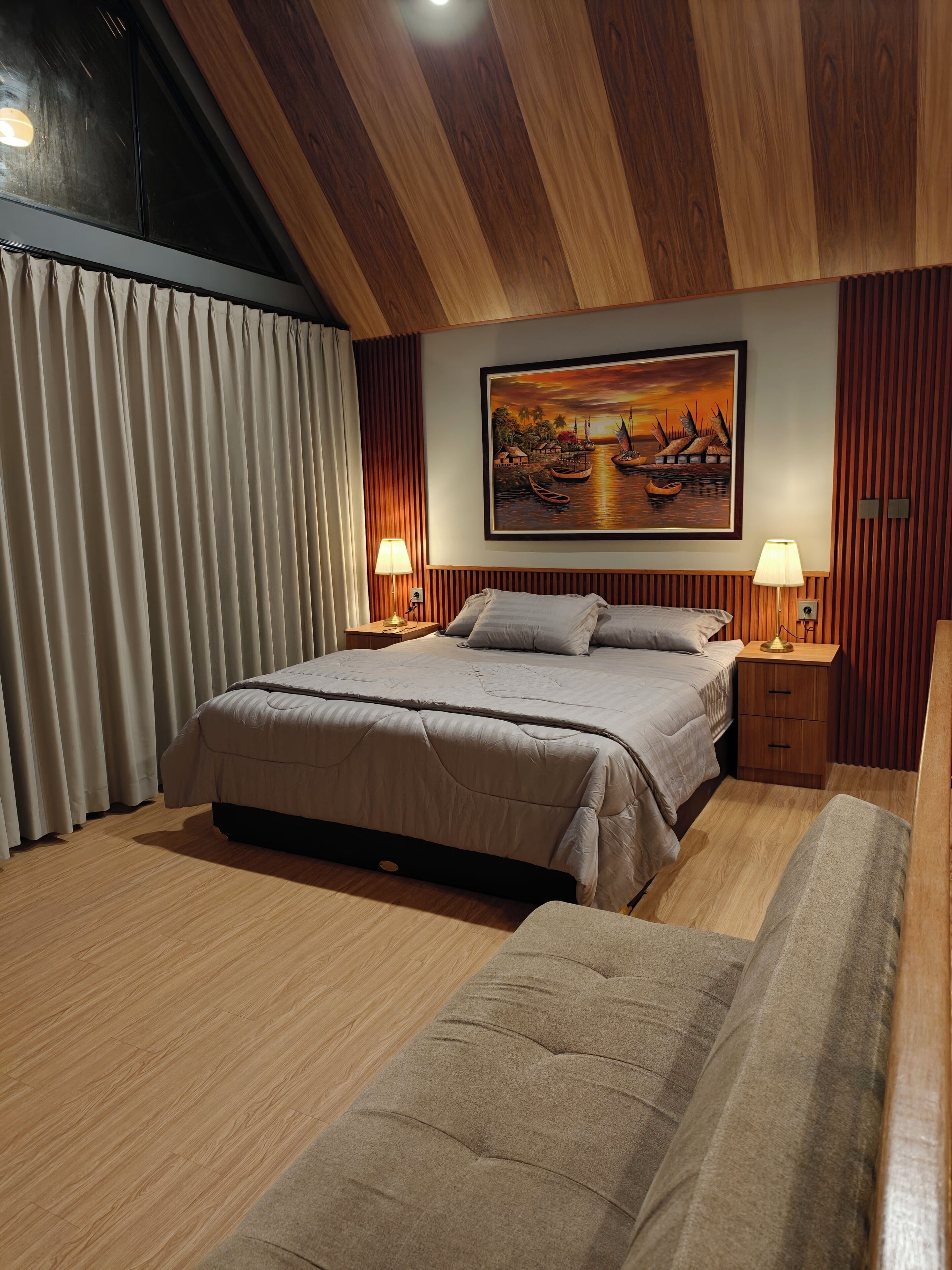 Un dormitorio con cama, mesitas de noche, un cuadro y una ventana con cortinas.