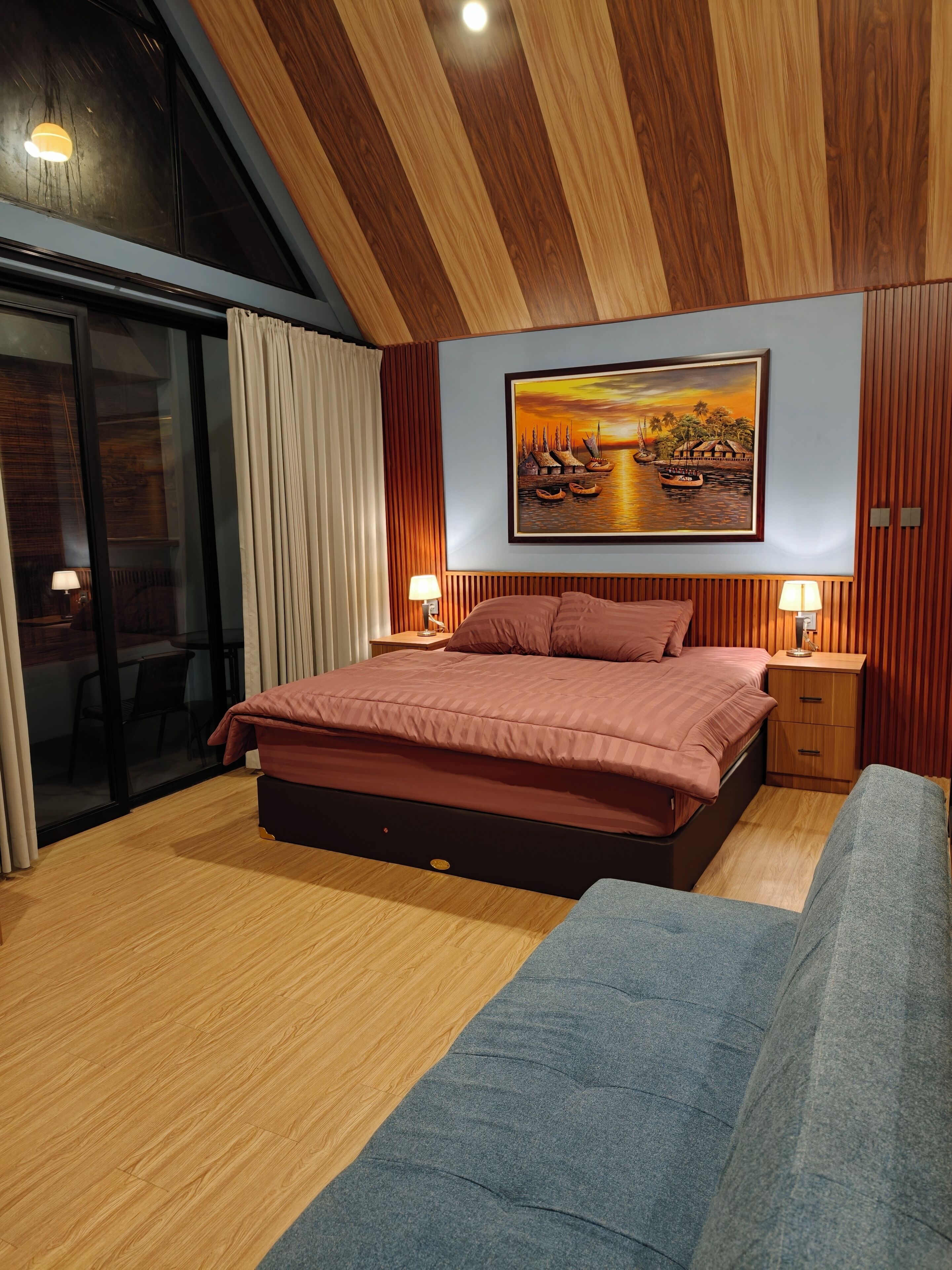 A modern bedroom with a large bed, a painting of a sunset scene, and a sliding glass door leading to an outdoor area.