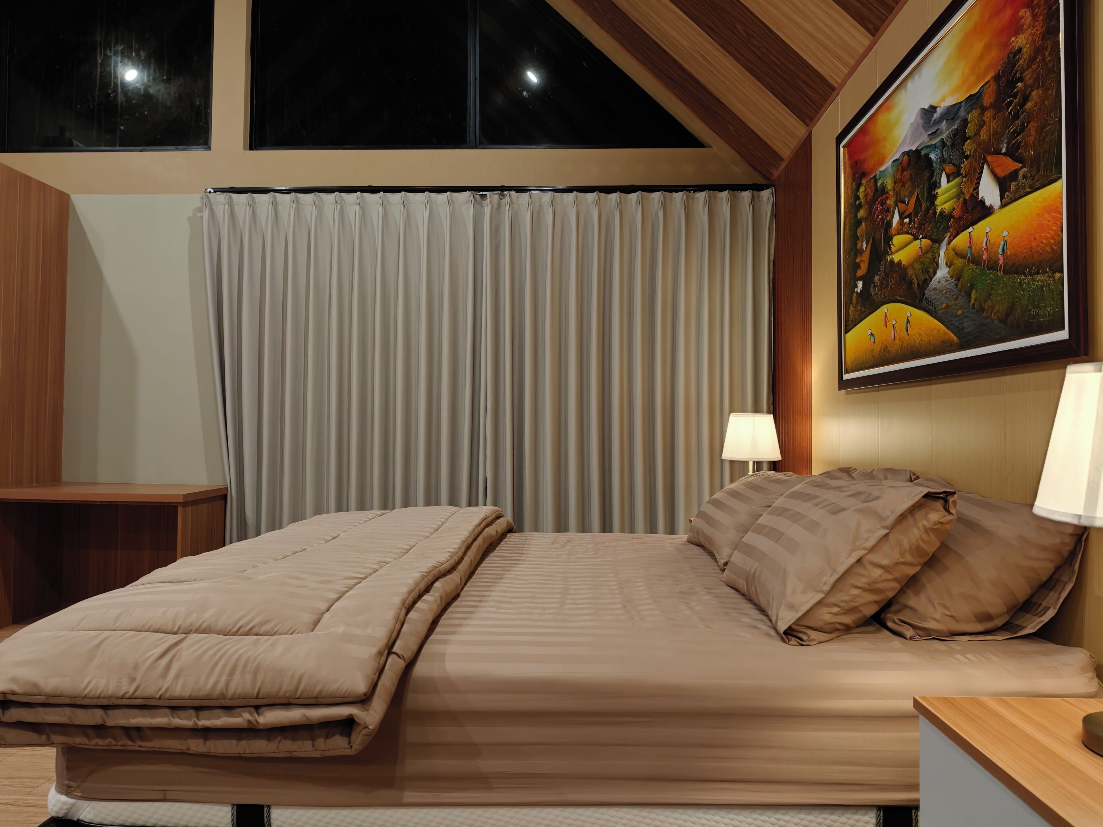 A bedroom with a bed, a nightstand, a lamp, a painting on the wall, and a window with curtains.