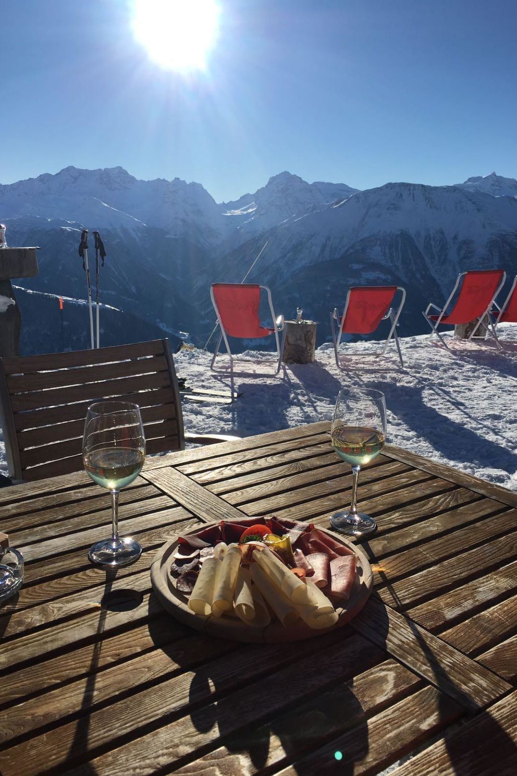 A wooden table with a plate of food and two glasses of wine, overlooking a snowy mountain landscape with red chairs and ski equipment.