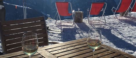 Una mesa de madera con un plato de comida y dos copas de vino, con vistas a un paisaje montañoso nevado y sillas rojas, además de equipo de esquí.
