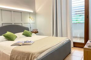 Ein ordentlich bezogenes Bett mit Kissen, ein Nachttisch mit Lampe und Blick auf einen Balkon mit Vorhängen.