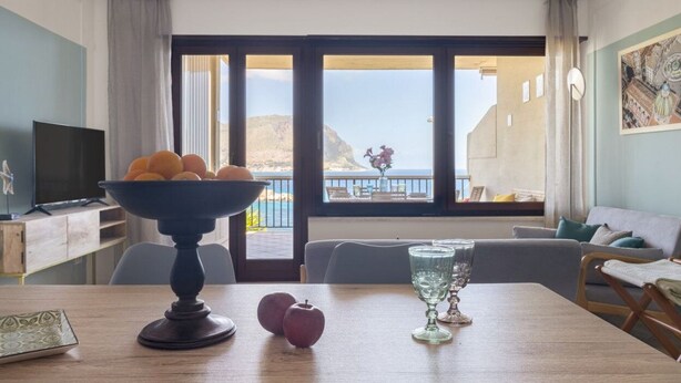 A room with a dining table, a bowl of fruit, a glass, and a television. - Mondello Bay - Addaura Apartments (Palermo)