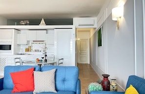 A living room with a blue sofa, a dining area with a table and chairs, white kitchen cabinets, and a hallway leading to a door. - Mondello Bay - Addaura Apartments (Palermo)