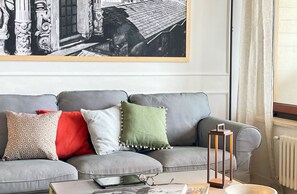 A grey sofa with patterned cushions, a framed black and white photograph above it, and a small table with a book and glasses. - Mondello Bay - Addaura Apartments (Palermo)