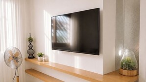 En moderne stue med et fladskærms-tv ophængt på væggen, træhylder, en gulvstandsf ventilator og dekorative planter.