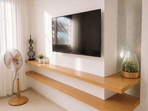 Ein modernes Wohnzimmer mit flachbildfernseher an der Wand, Holzregalen, einem Standventilator und dekorativen Pflanzen.