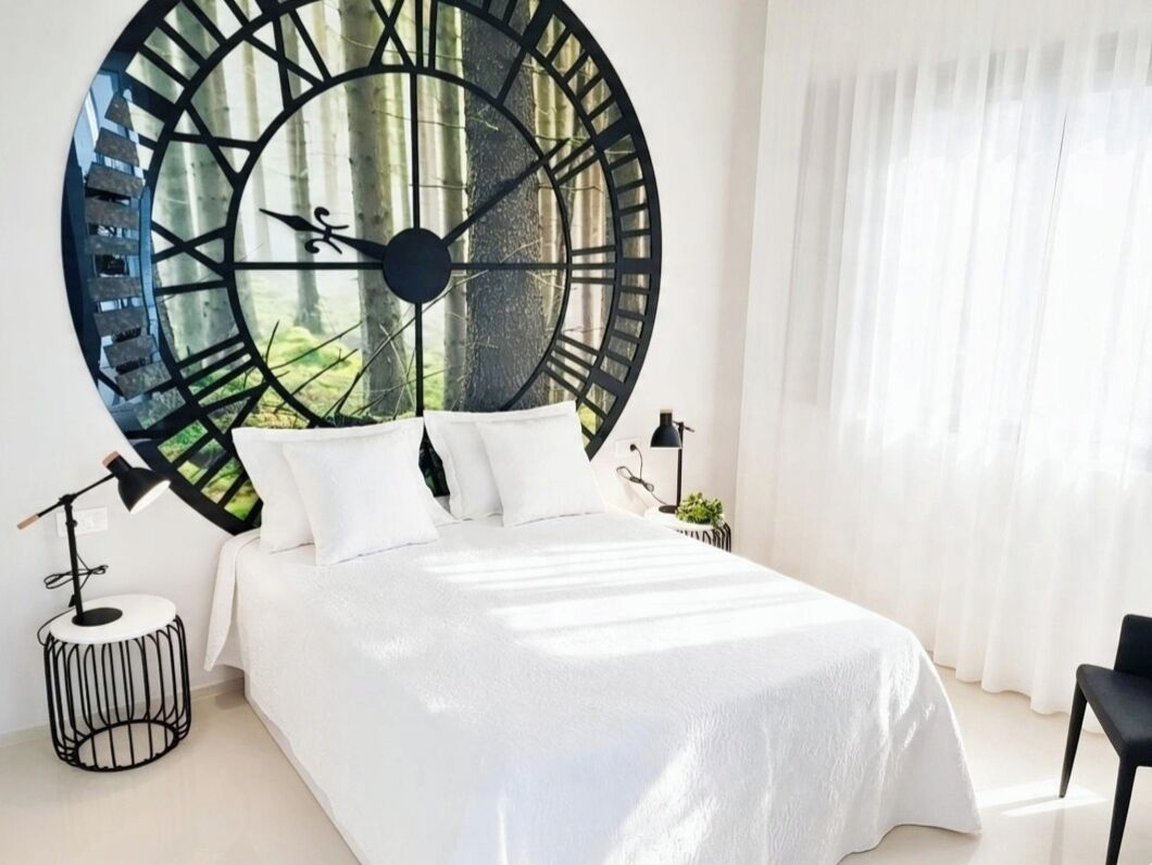 A bedroom with a large wall clock, a bed with white bedding, a side table, and a window with curtains.