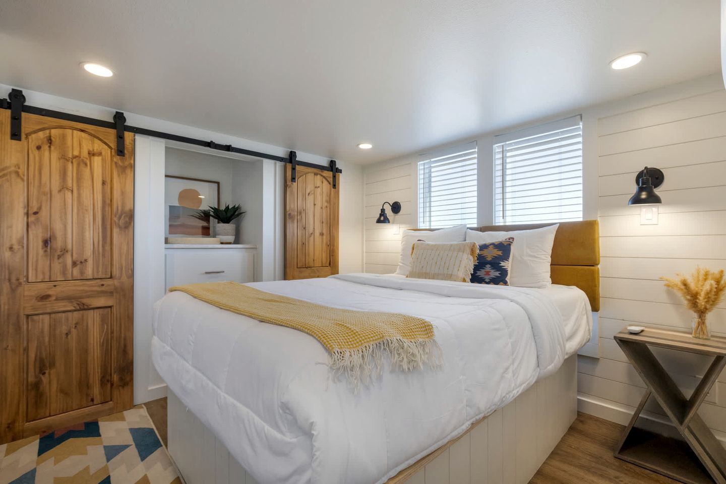 A bedroom with a bed, a wooden wardrobe, a bedside table with a lamp, and a window with blinds.