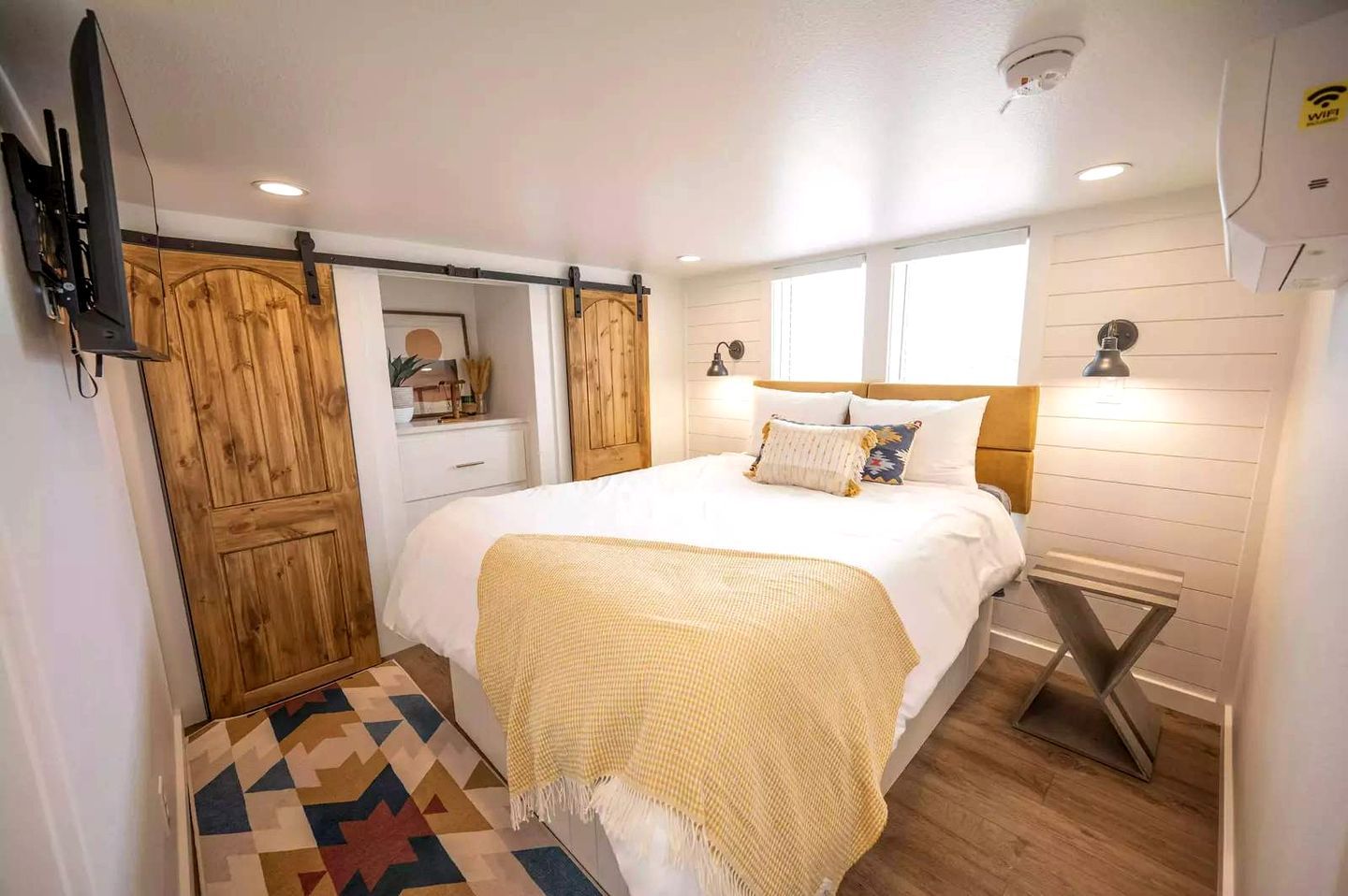 A bedroom with a bed, wooden wardrobe, bedside table, and a patterned rug.