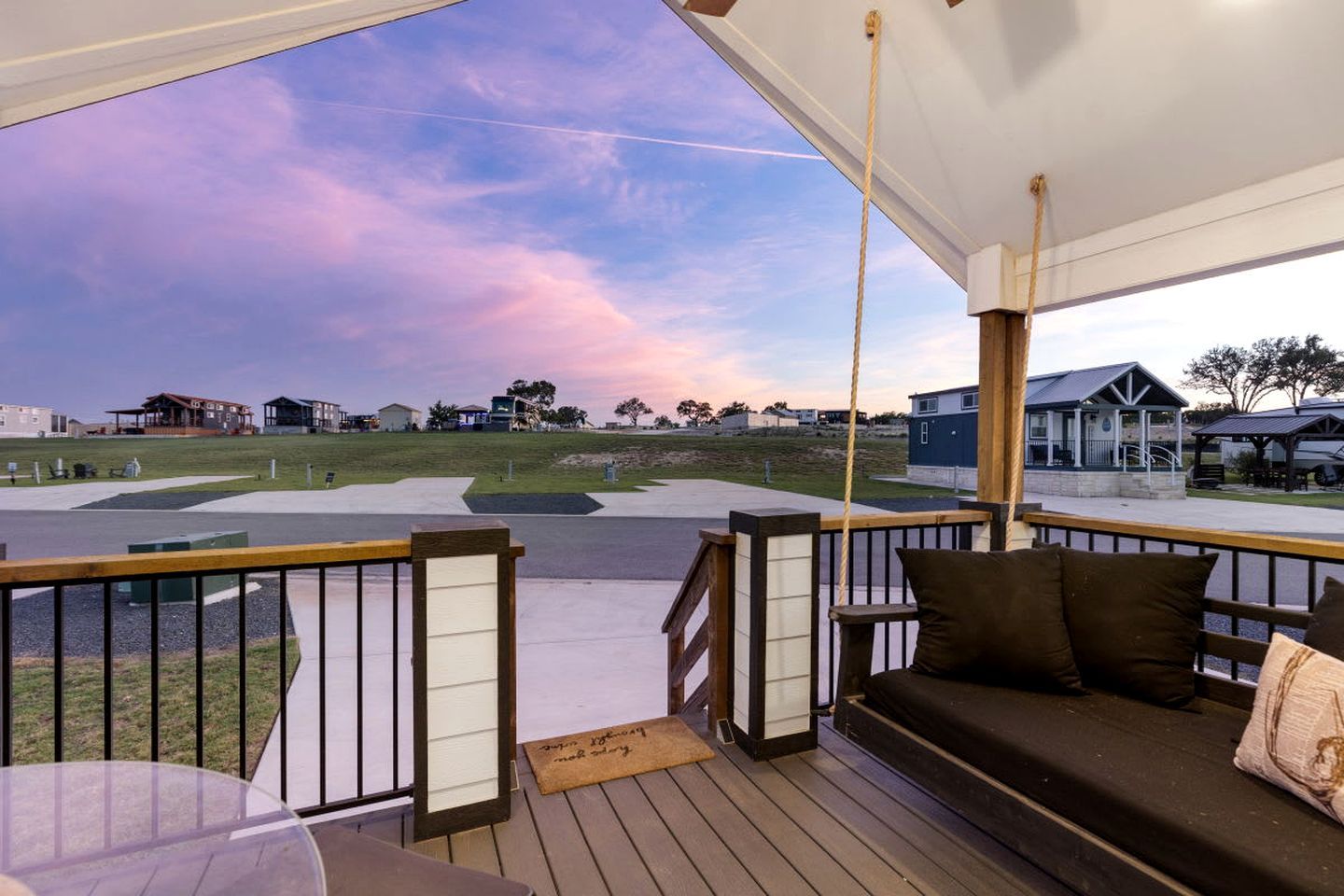A covered outdoor area with a wooden deck, a seating area with cushions, and a view of a grassy field and buildings in the distance.