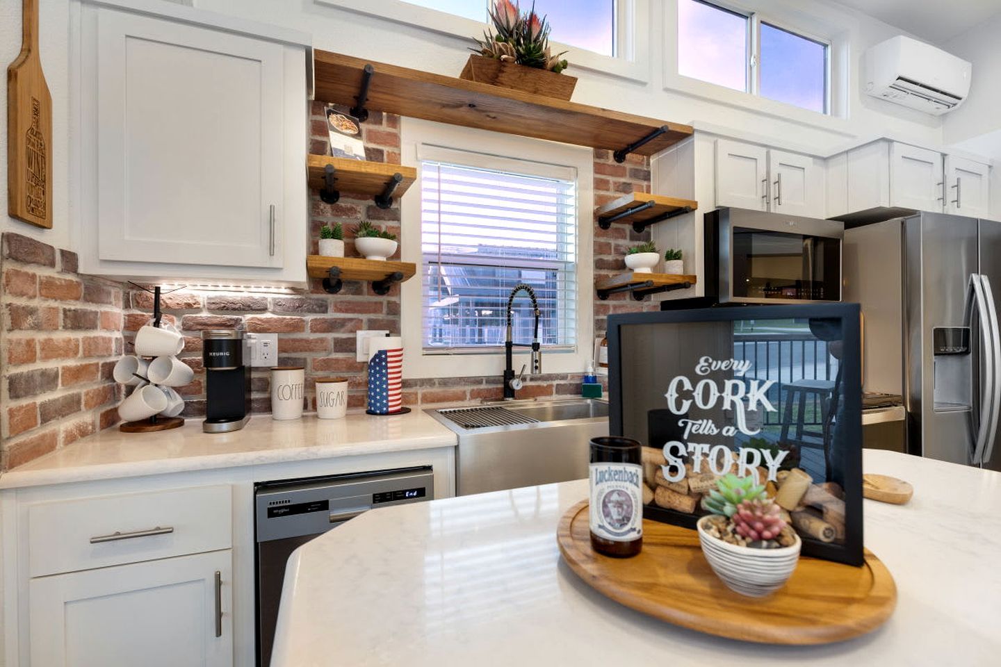 A kitchen with white cabinets, a brick accent wall, a coffee station, and a sign that reads 'Every Cork Tells a Story.'