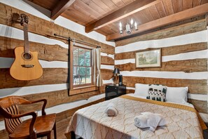 Habitació d'una cabana de fusta amb un llit, una guitarra, un escriptori i una finestra.
