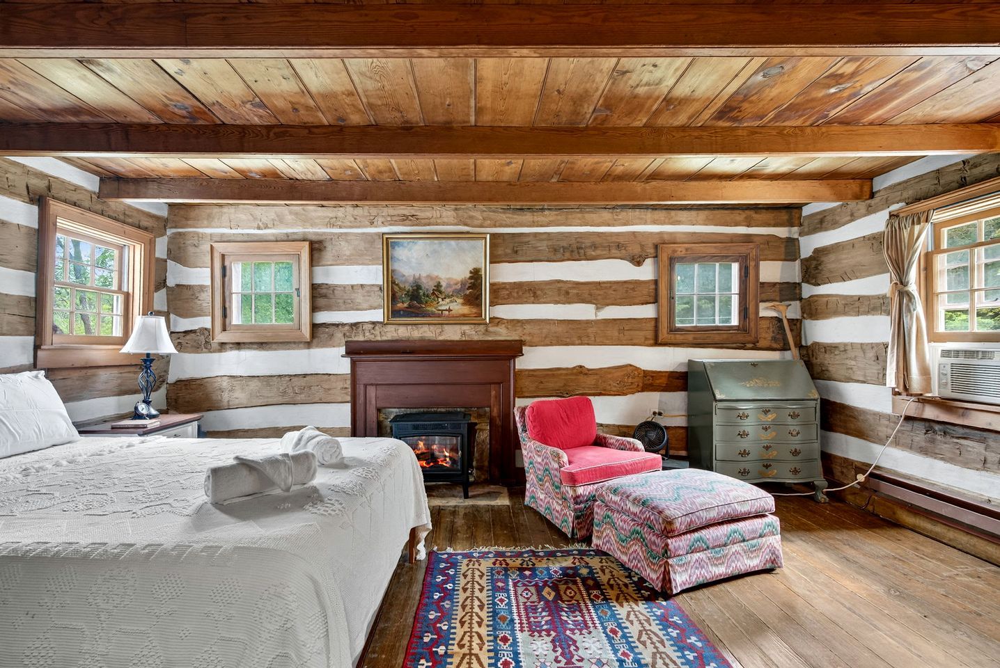 A log cabin bedroom with a bed, fireplace, chair, dresser, and two windows.
