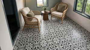 Une pièce dotée d’un sol à motifs, de deux fauteuils en osier, d’une petite table avec une lampe et de deux fenêtres surmontées d’arcs.