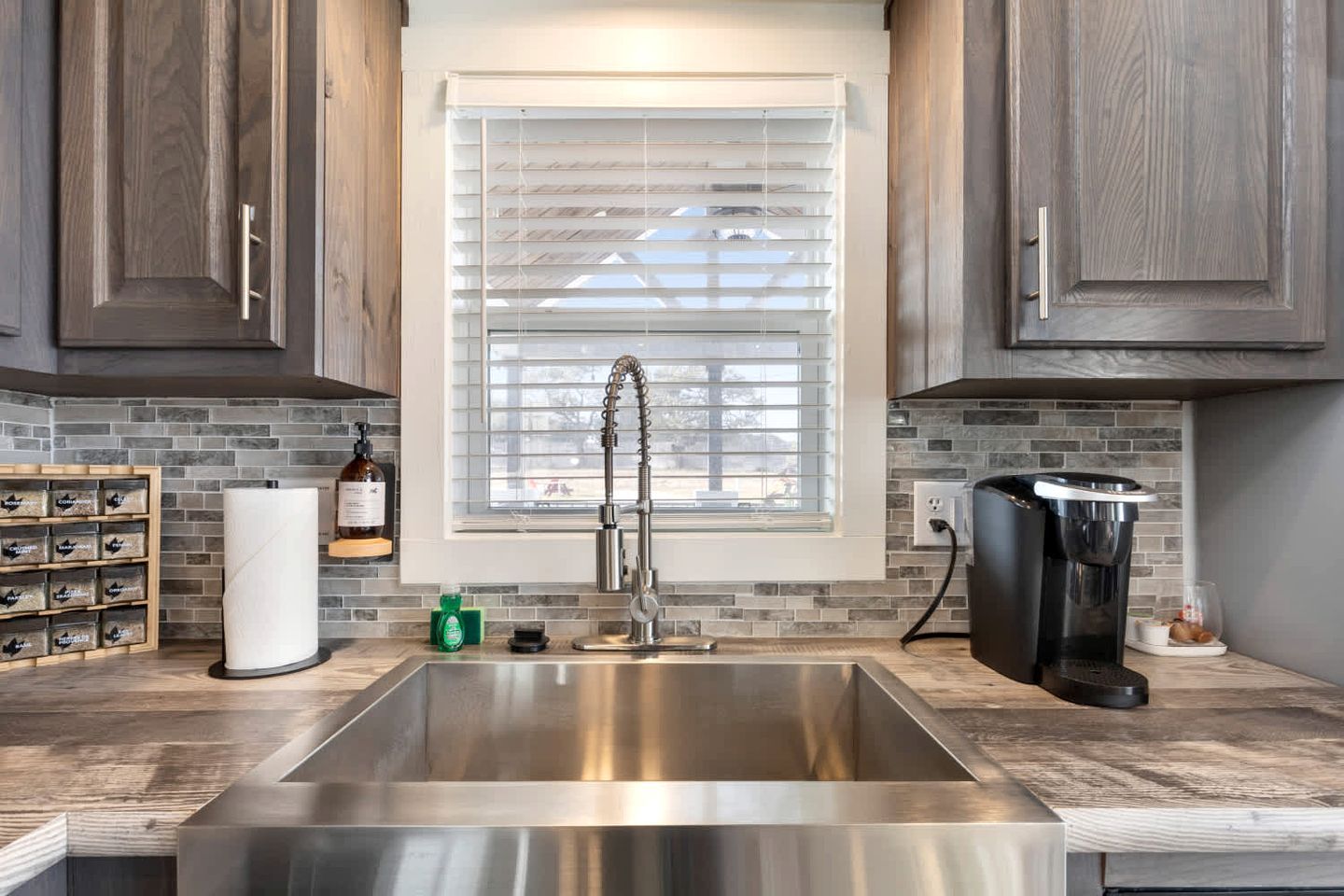 A modern kitchen with a stainless steel sink, wooden cabinets, a window with blinds, and a coffee maker.