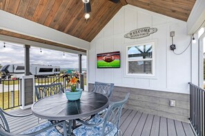 A covered patio area with a round table and chairs, a welcome sign, a television mounted on the wall, and a view of the outdoors. - Welcoming Tiny Home Getaway with Resort Amenities Near Fredericksburg, TX (Fredericksburg)