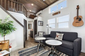 Un salon moderne avec un canapé gris, deux tables rondes, une plante en pot et une guitare accrochée au mur.