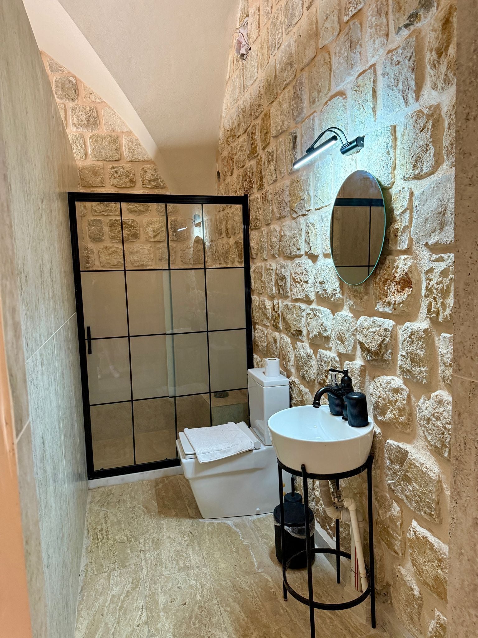 Un baño con pared de piedra, cabina de ducha de vidrio, lavabo blanco y un espejo.