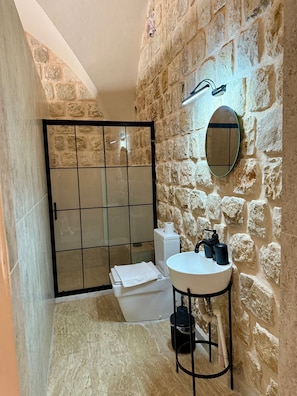 A bathroom with a stone wall, a glass shower enclosure, a white sink, and a mirror. - Evar Konağı (Mardin)