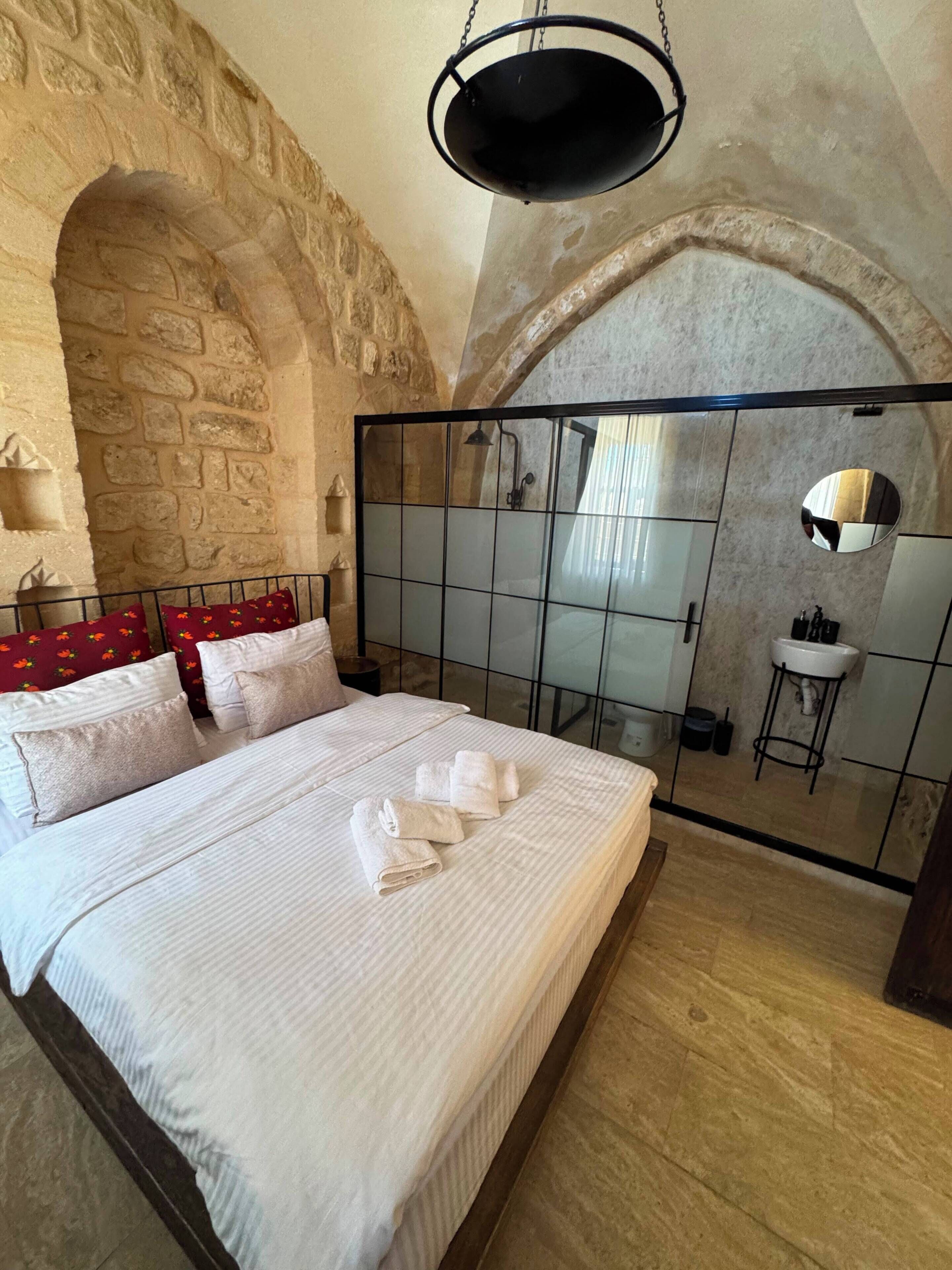 Un dormitorio con pared de piedra, una cama con ropa blanca, un colgante de luz y un box de ducha de vidrio.