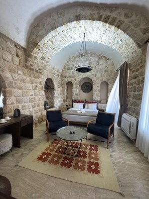 A stone-walled room with a bed, two armchairs, a small round table, and a hanging light fixture. - Evar Konağı (Mardin)