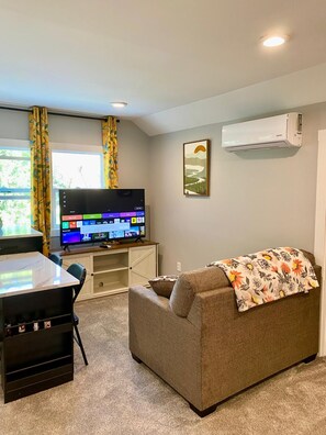 A living room with a flat-screen TV, a couch, a small table, and a white air conditioning unit on the wall. - Cute, Small, but Mighty! (Omaha)