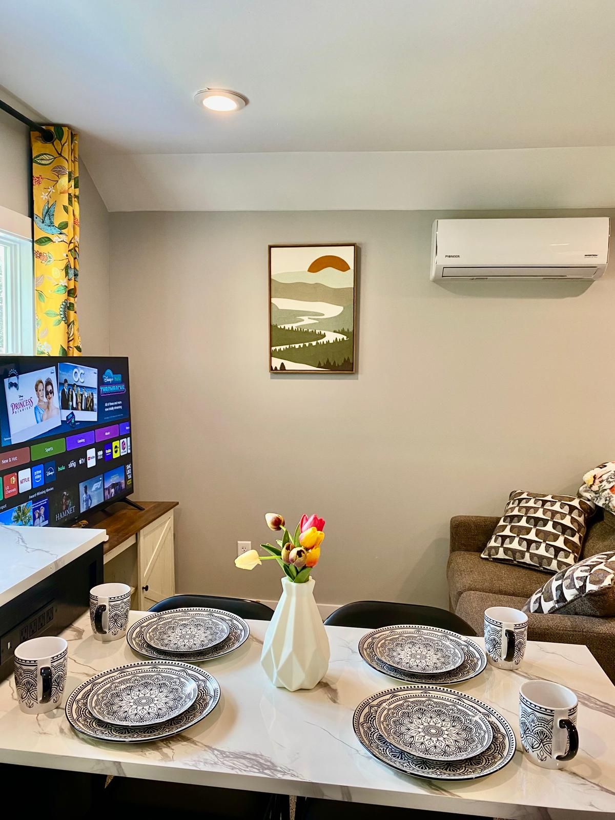 Un coin repas avec une table dressée pour quatre personnes, un vase rempli de fleurs, un téléviseur à écran plat diffusant un film et un appareil de climatisation fixé au mur.