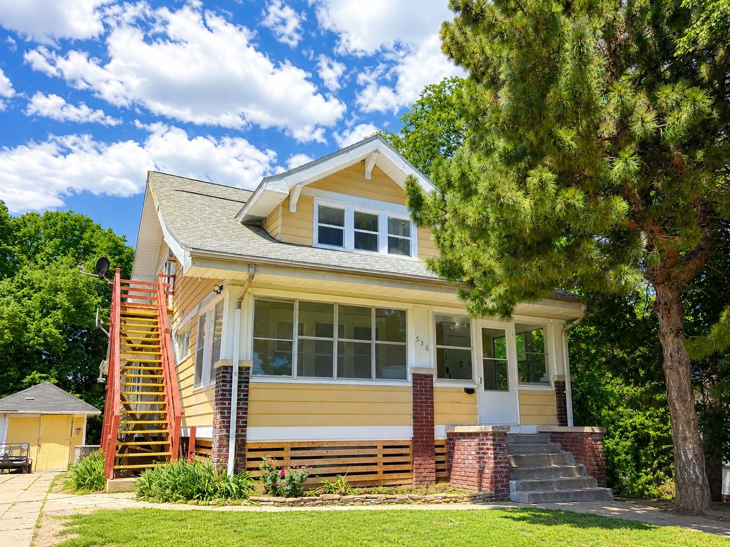 A two-story house with a front porch, a red staircase, and a large tree in the background.