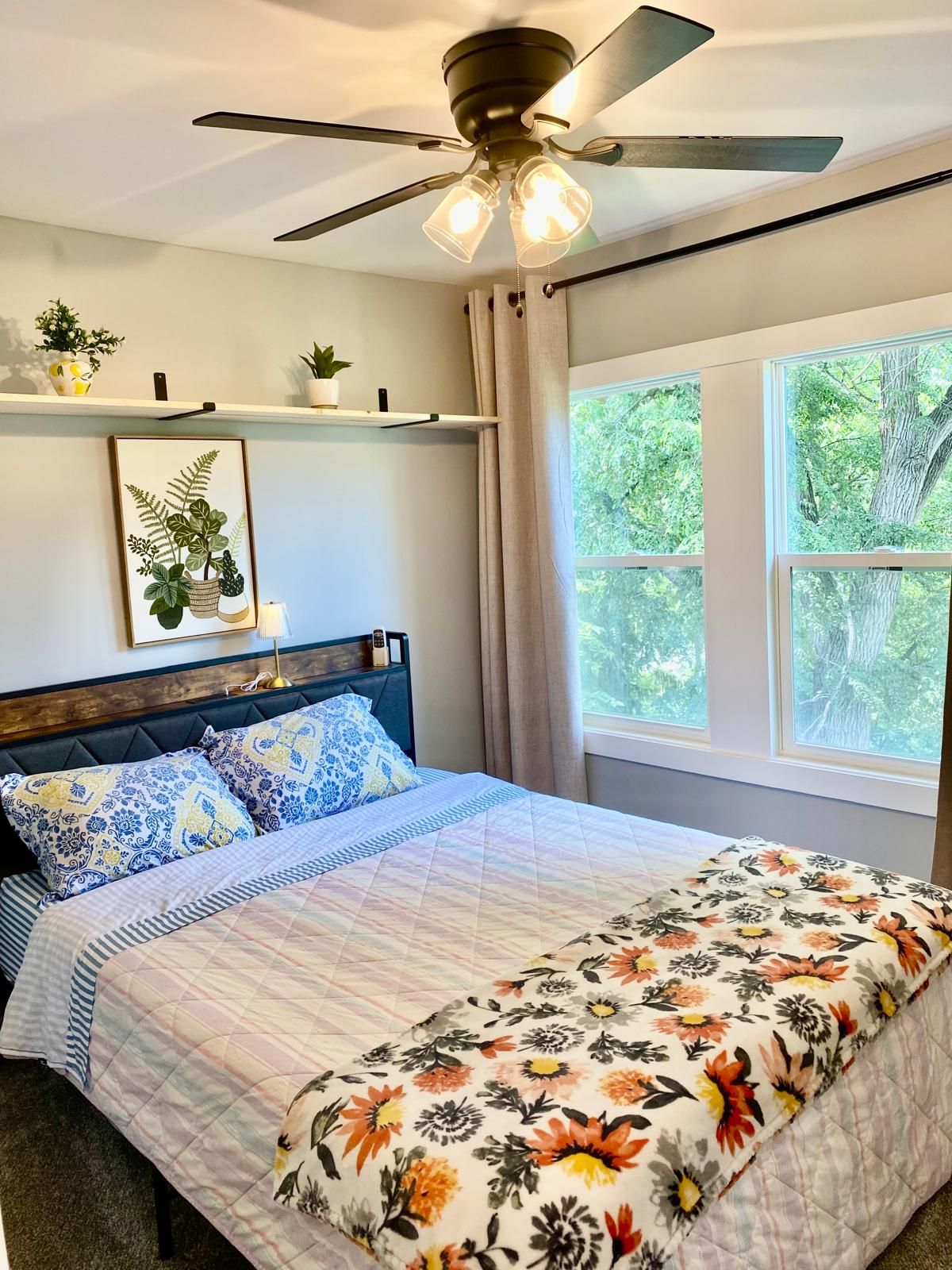 A bedroom with a bed, a ceiling fan, a window with a view of trees, and a shelf with plants.