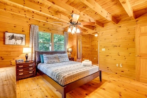 A wooden cabin bedroom with a bed, nightstands, a ceiling fan, and a window with curtains.