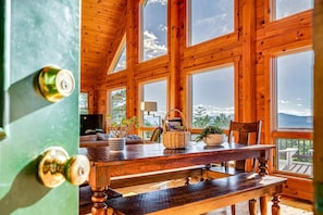 A wooden interior with a dining table, chairs, a TV, and large windows offering a view of the outdoors.