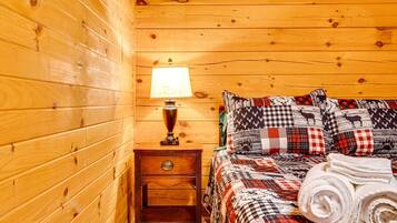 A wooden cabin room with a bed, a nightstand, a lamp, and a rolled towel.