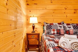 A wooden cabin room with a bed, a nightstand, a lamp, and a rolled towel.