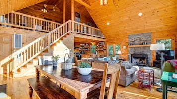 A spacious wooden interior with a central dining area, a fireplace, and a staircase leading to an upper level.